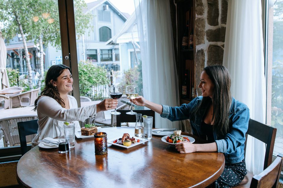 Two girlfriends clink their wine glasses together over a table of fresh greek dishes made at Tholos Restaurant in Blue Mountain Village.