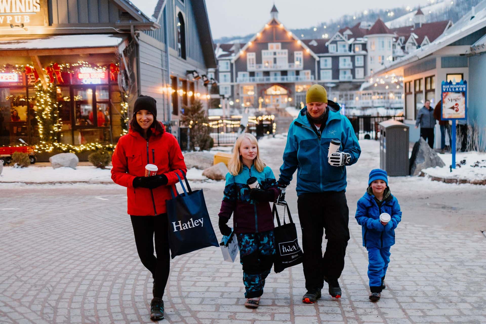 Family of four, each holding a hot beverage and a shopping bag, strolling through the festively lit pedestrian Village.