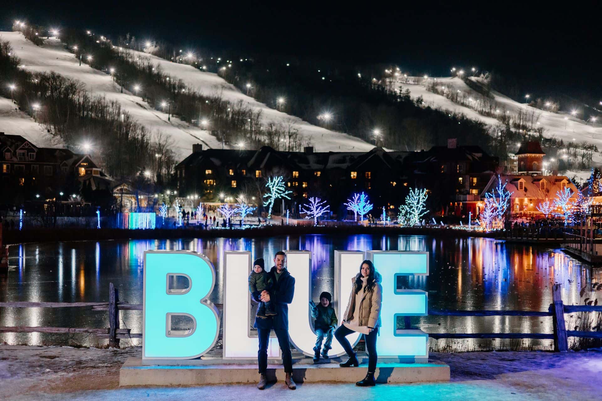 Family of four taking a group photo in front of the lit up BLUE sign, with the Village and Mountain lit up behind them.