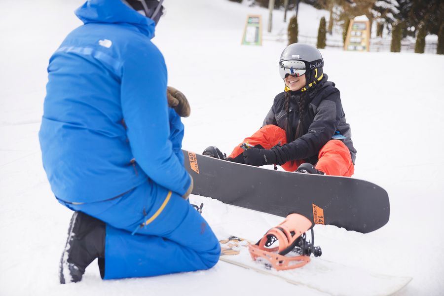 Woman doing up her snowboard bindings, while an instructor guides her.
